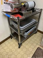 Full view of NSF stainless steel utility cart with three shelves and four gray plastic bus trays on middle and bottom shelves, pictured in kitchen area.