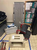 Full view of two metal folding steps, rolling mechanic stool with black padded seat and red frame, and beige plastic step stool