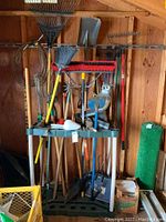 Wide shot of the full garden tool rack and various tools leaning on shed wall, including owl decoy, rakes, broom, hoe.
