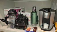 Vitamix blender base and container next to Stanley thermos and Keurig machine with paperwork and cup.