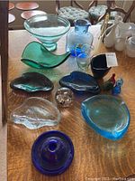 View of all 14 art glass pieces on a wooden table including bowls, pitchers, decorative bottles, and paperweight.