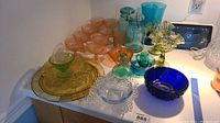 Overall view of the 25 pieces of colored glassware showing various colors, shapes and sizes including peach goblets, amber plate, blue vase and bowls.