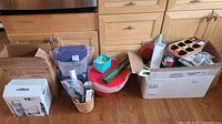 Wide view of all kitchen and serving items arranged on hardwood floor, including boxed Pampered Chef Chillzanne Platter, silicone molds, glass bowls, red storage container, small utensils in basket, and various boxes.