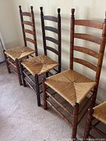 Five antique wooden ladder back chairs with rush seats lined up on carpeted floor showing their overall structure, wood finish, and woven seats.
