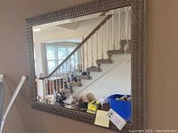 Full view of the rectangular silver framed mirror hanging on the wall, reflecting a staircase and window.