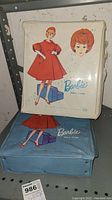Two vintage Barbie doll cases stacked, white on top and blue below, each with illustrated cover of a red-haired Barbie in a red dress holding a blue suitcase.