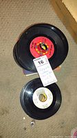 Top-down view of two 45 RPM vinyl records, showing labels and condition with a lot tag attached indicating 'Records'