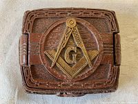 Top view of the wooden box lid showing the carved Masonic square and compass emblem with a 'G' in the center, detailed carvings on the border, and hinge on the right side.