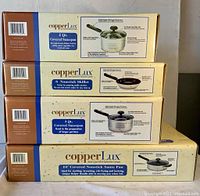 Four stacked cardboard boxes of CopperLux cookware showing box labeling including 2 qt and 3 qt covered saucepans, 8" nonstick skillet, and 10" covered nonstick sauté pan.