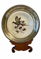 Front view of the silver-rimmed dish showing the hand-colored pheasant design and silver rim tarnish on a wooden stand.