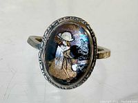 Close-up view of the painted glass ring showing the detailed painting of the girl in white hat holding a black dog, with visible reflective glass surface and textured metal bezel.