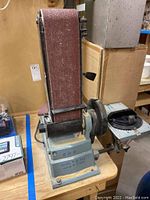 Full view front angle of the Delta belt sander attached to a wooden table, sanding belt running vertically.