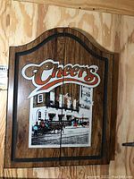 Front view of the wooden wall-mounted dart board cabinet showing Cheers logo and bar image