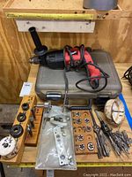 Full view of Milwaukee hammer drill placed on grey metal carrying case along with wooden holders of dies and taps, assorted screws, hole saw, and drill bits.