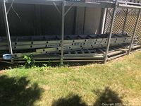 Three aluminum extension ladders stacked on the ground outside under a canopy, viewed from the side.