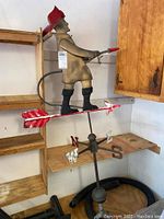 Left side profile of the painted fireman weathervane showing figure, red helmet, hose, and arrow with directional letters.