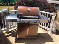 Front view of bronze Weber gas grill on deck, showing closed lid, control knobs, and side shelves.