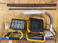 Two yellow jobsite lamps, a white flexible desk lamp, and two saw blades on wooden surface