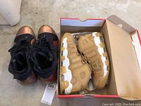 Two pairs of Nike sneakers, one brown/tan pair in box, one black with copper toe caps outside the box.