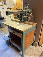 Full view of the rip saw mounted on metal and wood stand in basement setting with visible drawer and open storage underneath.