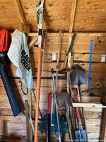 Photo showing a variety of garden tools including hedge trimmer pole, rakes, shovels, and pickaxe leaning against a wooden wall inside a shed.
