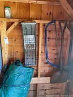 Photo showing green tarp, Havahart metal animal trap hanging on wall, and garden tools including shovels and bow saw.