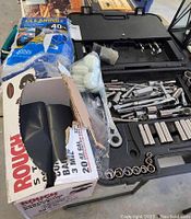 Socket wrench set with various tools and extensions organized inside a black plastic case with a partially opened Rough Tough contractor bag box and disposable gloves beside it.