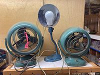 Overview of two vintage Vornado desk fans and a blue desk lamp on a shelf.