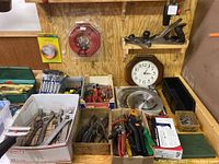 Wide angle image showing multiple hand tools in open boxes and on a wooden table: adjustable wrenches, pliers, screwdrivers, hardware boxes, a clock on the wall, and saw blades in the foreground.