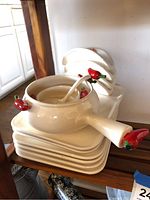 Stack of cream-colored ceramic soup bowls with chili pepper shaped handles and matching spoons, stacked on square plates with rounded edges, set on a wooden shelf.