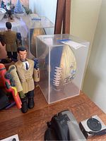 Inspector Gadget toy standing next to the Disney football enclosed in a clear display case on a wooden surface.