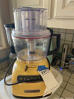 Front view of yellow KitchenAid food processor showing the feed tube, clear processing bowl, and base with control panel and brand logo.