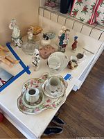 Porcelain dishes with floral patterns, porcelain figurines, glassware, and small decorative items arranged on a white table.