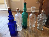 Five vintage glass decanters arranged on wooden floor by door, showing different shapes, embossing, and colors with cork stoppers.