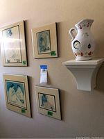 Wall view showing four framed plaques and a white ceramic pitcher on a wall shelf, providing overview of items in lot.