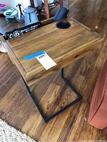 Top and angled view of the wooden side table showing wood grain finish, built-in cup holder, and metal cantilever base sitting on wooden floor.