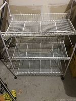 3 tier metal shelving table with mesh shelves, white painted metal, stored indoors, on caster wheels.
