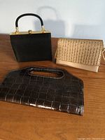 Photo of three clutch purses on wooden surface: black box clutch with gold hardware, beige woven-texture clutch, dark brown croc-embossed clutch