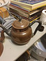 Copper teapot with black handle and hinge lid seen on tabletop with stacked magazines in background.