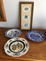 Three decorative plates on wooden surface including a round windmill floral plate, a blue white scenic plate, and a blue white octagonal plate. Next to a gold-framed artwork with three blue teacup drawings.