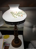 Hurricane lamp with carved wood base and frosted white glass shade painted with green leaves and orange flowers, photographed on a white surface next to a chair and magazine.