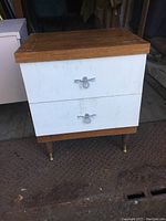 Front view of mid-century modern bedroom end table showing white drawer fronts, metal loop handles, wood top and base, brass-tipped legs