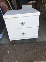 Front view of MCM bedroom end table showing two drawers with metal loop handles, white finish, and tapered wooden legs with brass feet caps. Condition visible with wear and scuff marks.