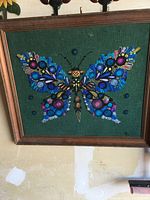 Full view of framed needlepoint butterfly on green fabric, showing the entire butterfly centered and wooden frame edges.