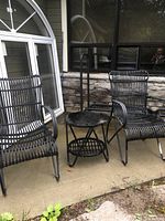 Two black outdoor armchairs and small round table made of metal frames with rattan-style weave, placed on a patio against a window and stone wall.