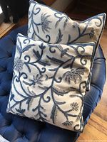Two blue and white square decorative pillows with raised floral and vine embroidery on a white base, placed on a dark blue tufted chair.