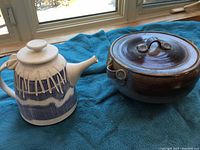 Photo showing a pottery tea pot with blue drip glaze decoration and a covered casserole dish with a brown glaze placed on a blue cloth by the window.