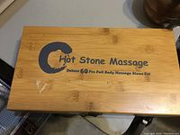 Closed bamboo box with logo and product name on top reading 'Hot Stone Massage Deluxe 60 Pcs Full Body Massage Stone Set'.