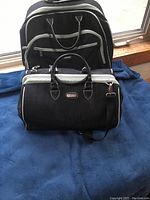Two black suitcases placed on a blue surface in front of a window, showing the front side of both with visible zippered compartments and handles.