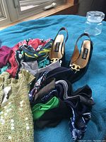 Lot arranged on blue fabric: one pair of black patent leather slingback shoes with gold emblem, colorful scarves, and folded tops in assorted colors and patterns
